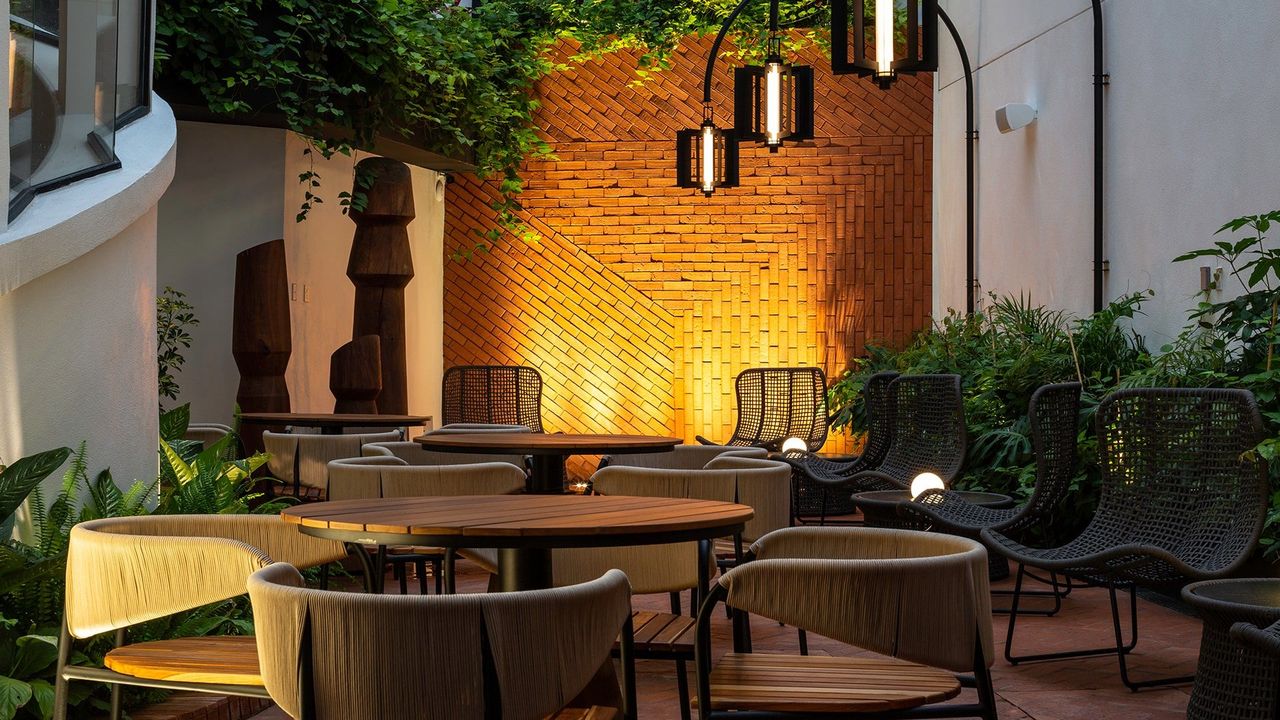 Patio del Hotel Campos Polanco de noche: mesas de madera, sillas tejidas, plantas y lámparas colgantes frente a un muro de ladrillo iluminado.